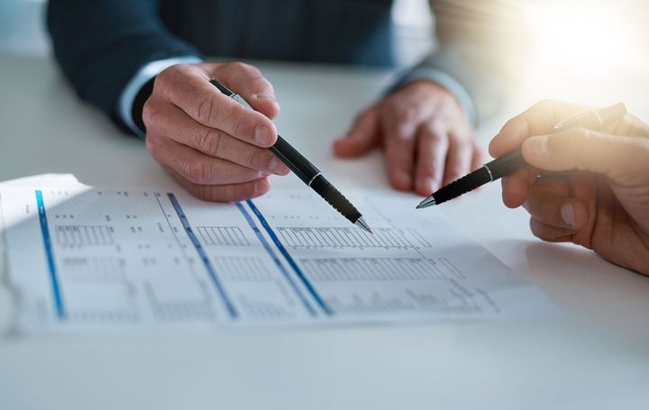 Two people in suits reviewing paperwork, pointing pens at document on white table.