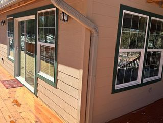 Exterior corner of a house with tan siding, green-trimmed windows, and a wooden deck in sunlight