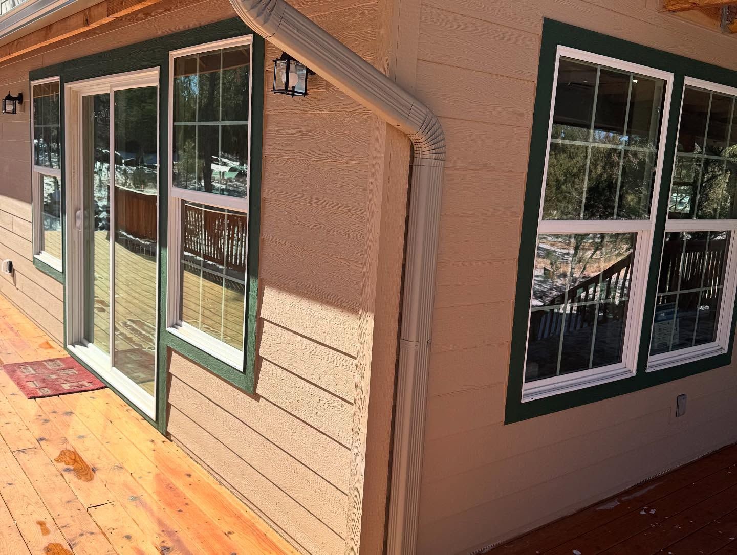 Exterior corner of a tan house with green-trimmed windows and a wooden deck in sunlight