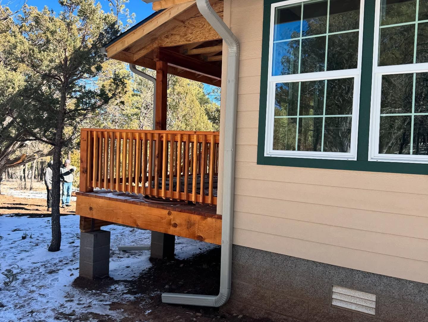 Snowy porch with tan house siding, white-trimmed window, wooden railing, and a downspout on concrete blocks