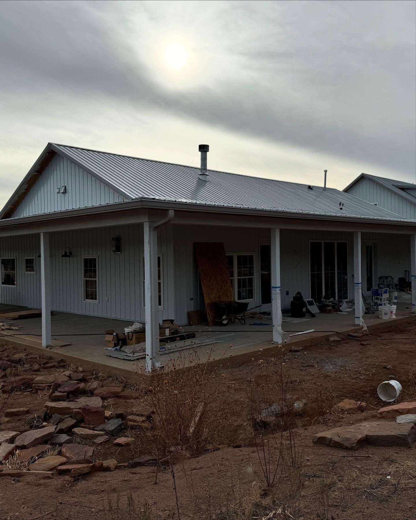 White rural house with a metal roof and porch, surrounded by bare ground under a cloudy sky