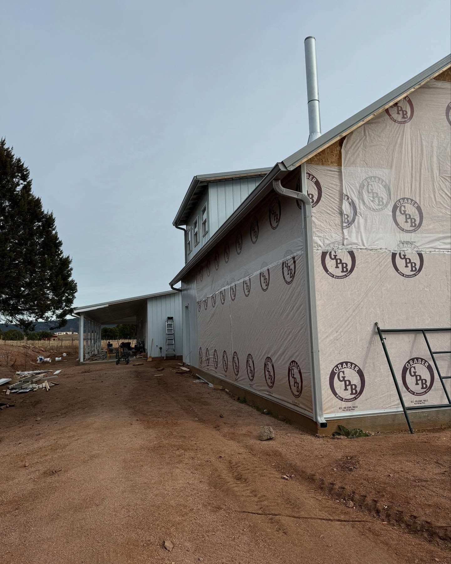 Partially finished house with gray siding, wrap and chimney on a dirt lot under overcast skies