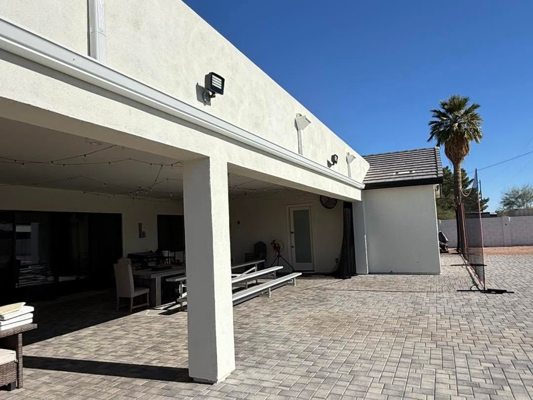 Covered patio with white columns, seating, and a paved courtyard beside a building under a clear blue sky
