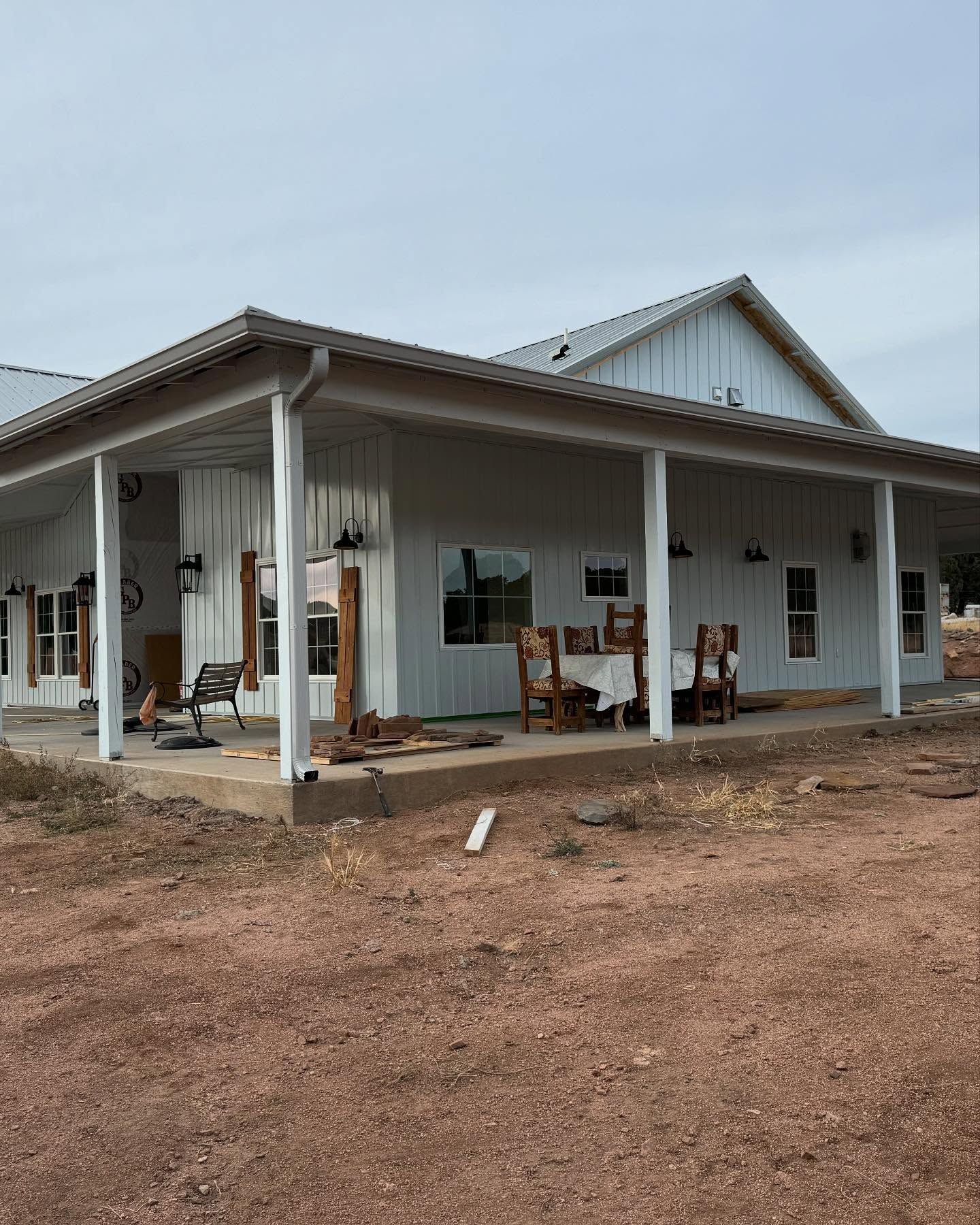 White farmhouse-style building with porch chairs on a dirt lot under an overcast sky