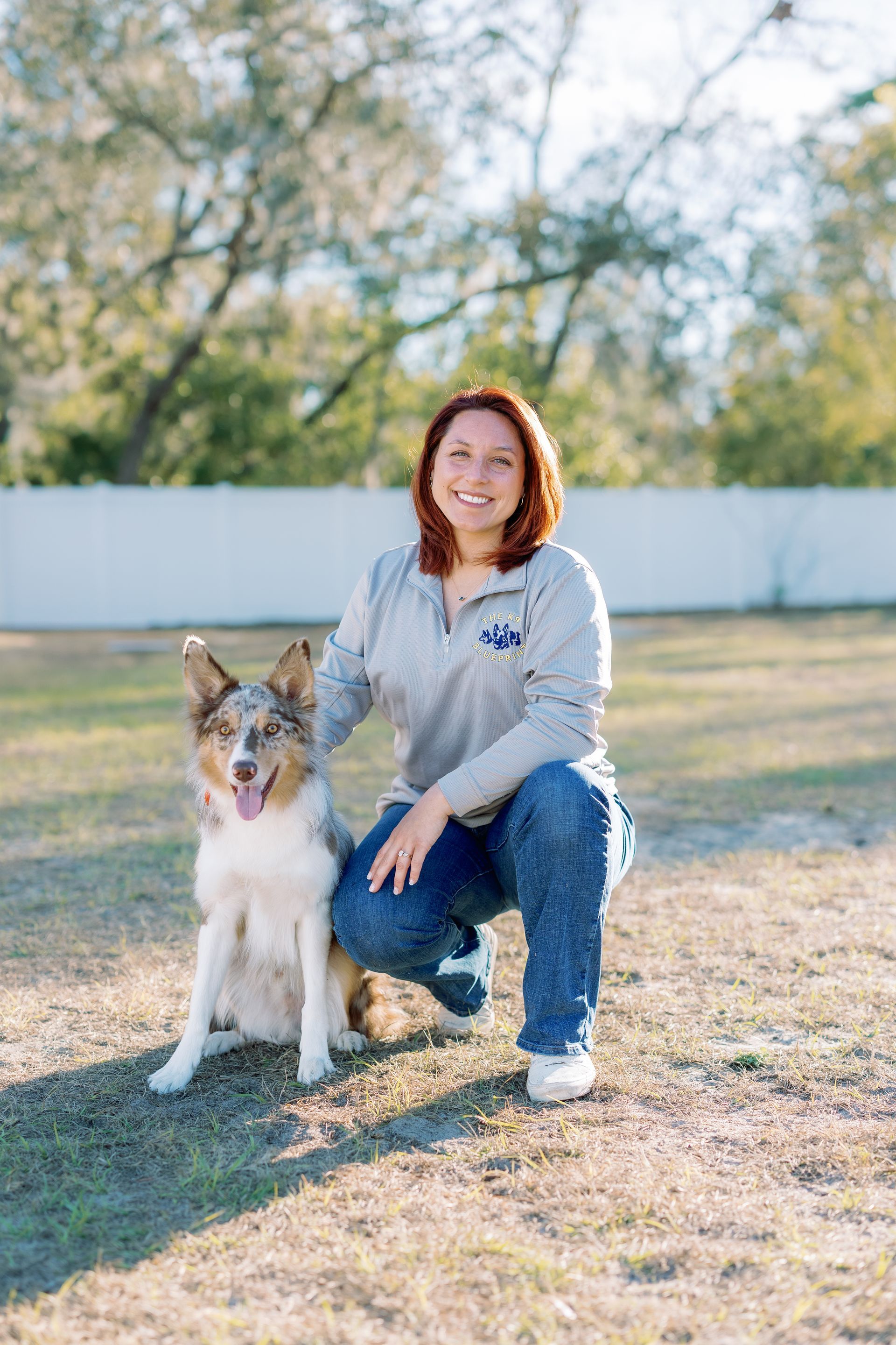Woman crouching beside a dog on a grassy field, with trees and a white fence in the background.