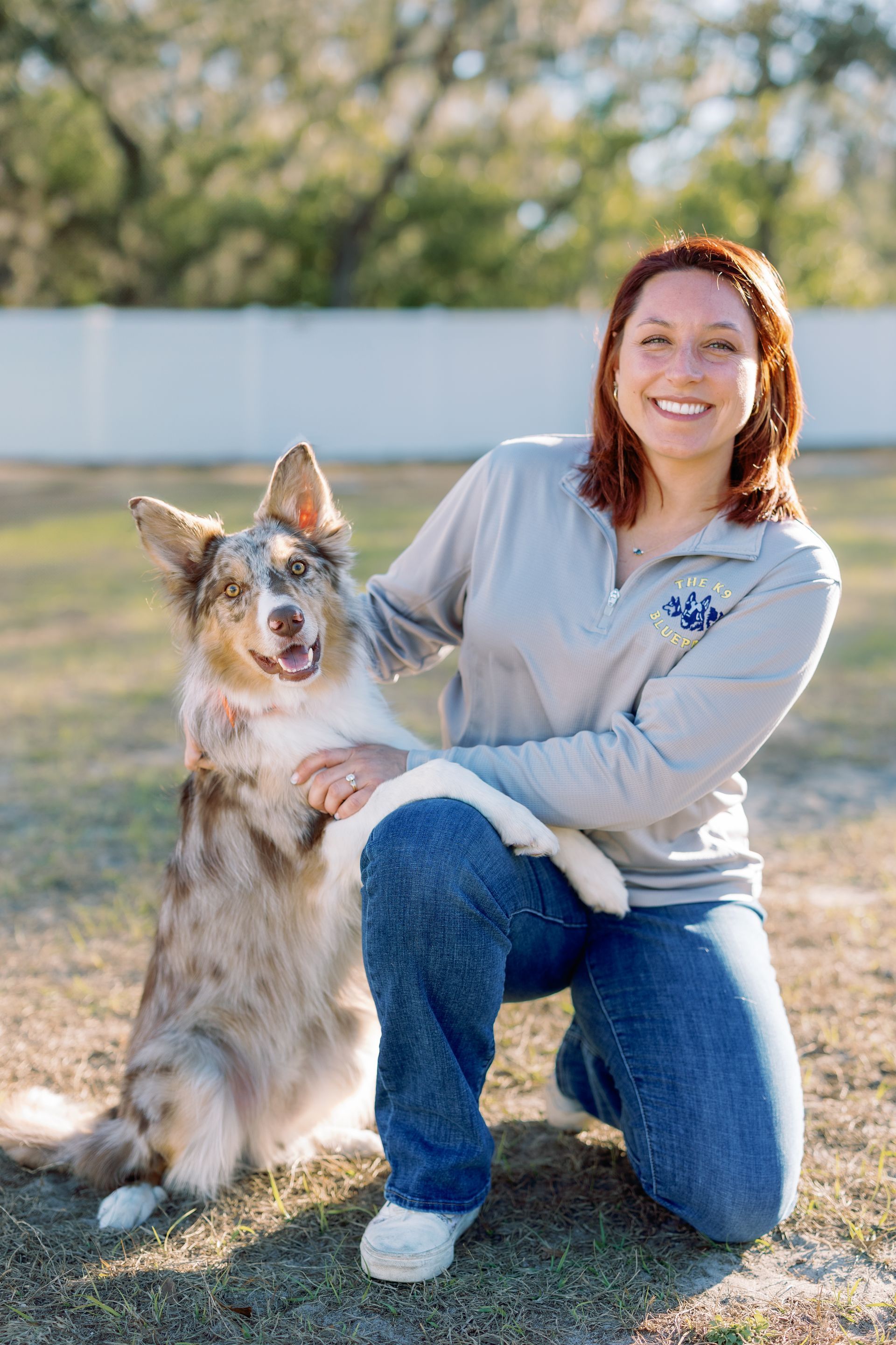 Person kneeling outdoors with a tan dog, both smiling in a sunny yard