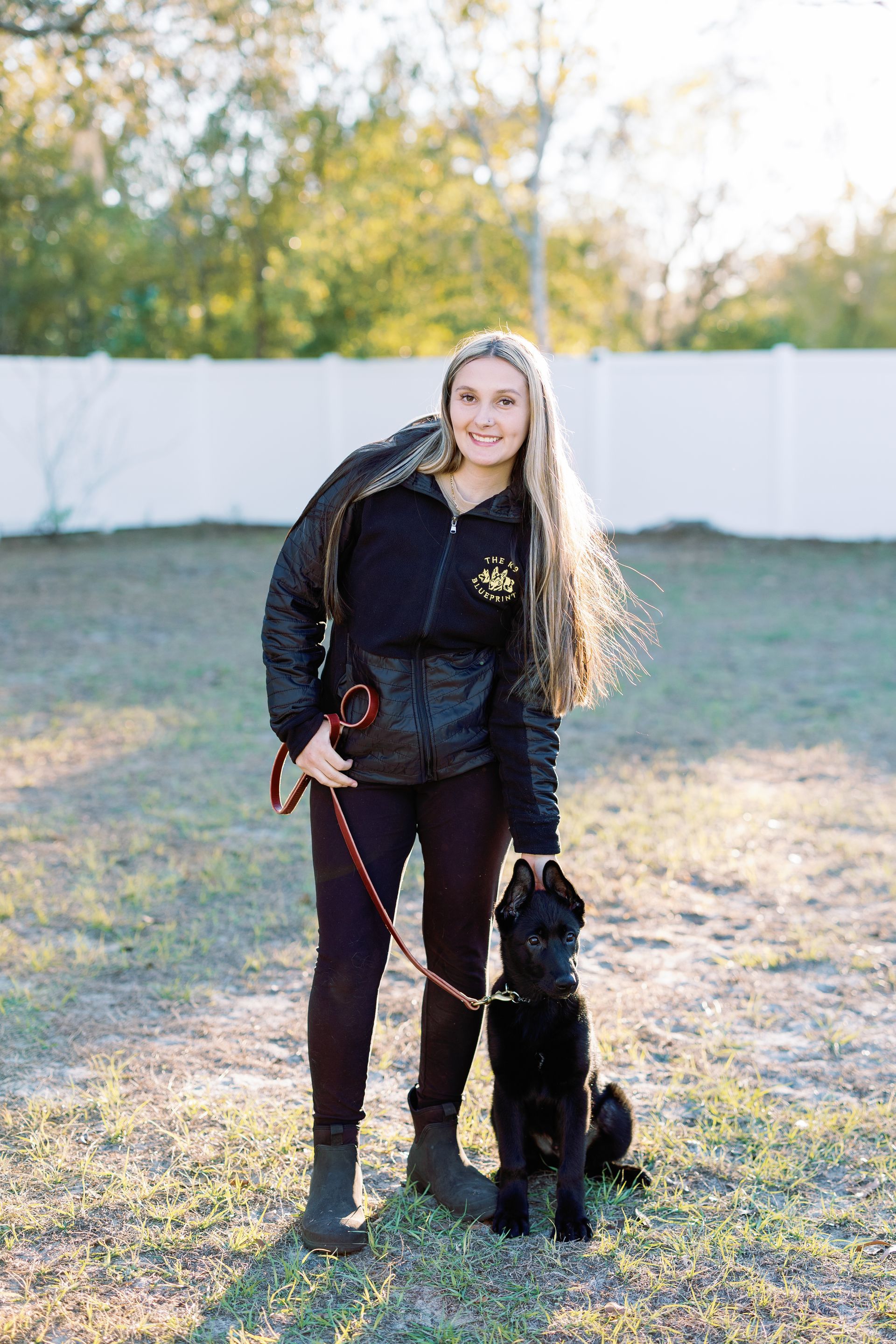 Woman in a black jacket standing outdoors with a black dog on a leash in a grassy yard.