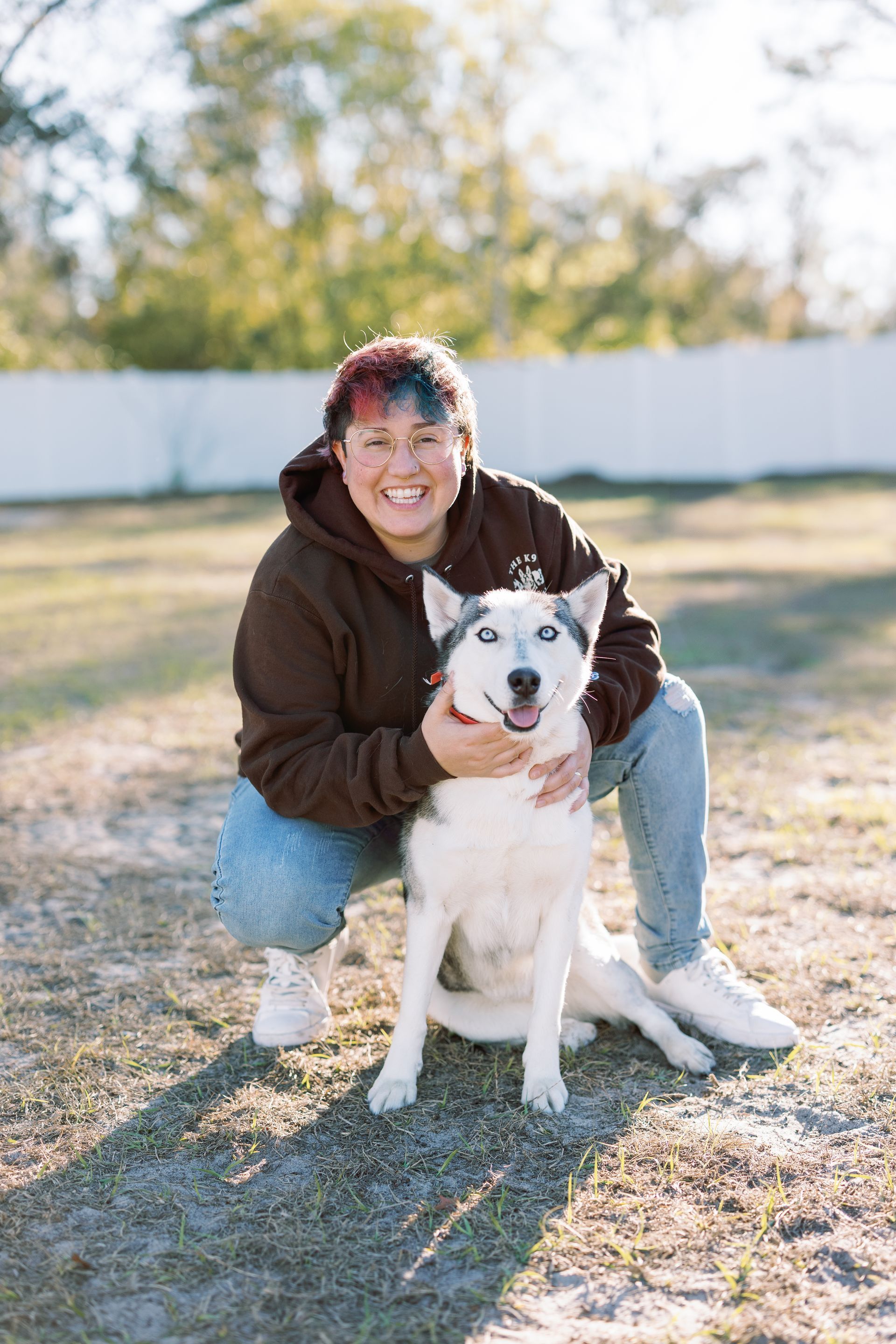 Person crouching beside a white dog in a sunny yard, both facing the camera and smiling.