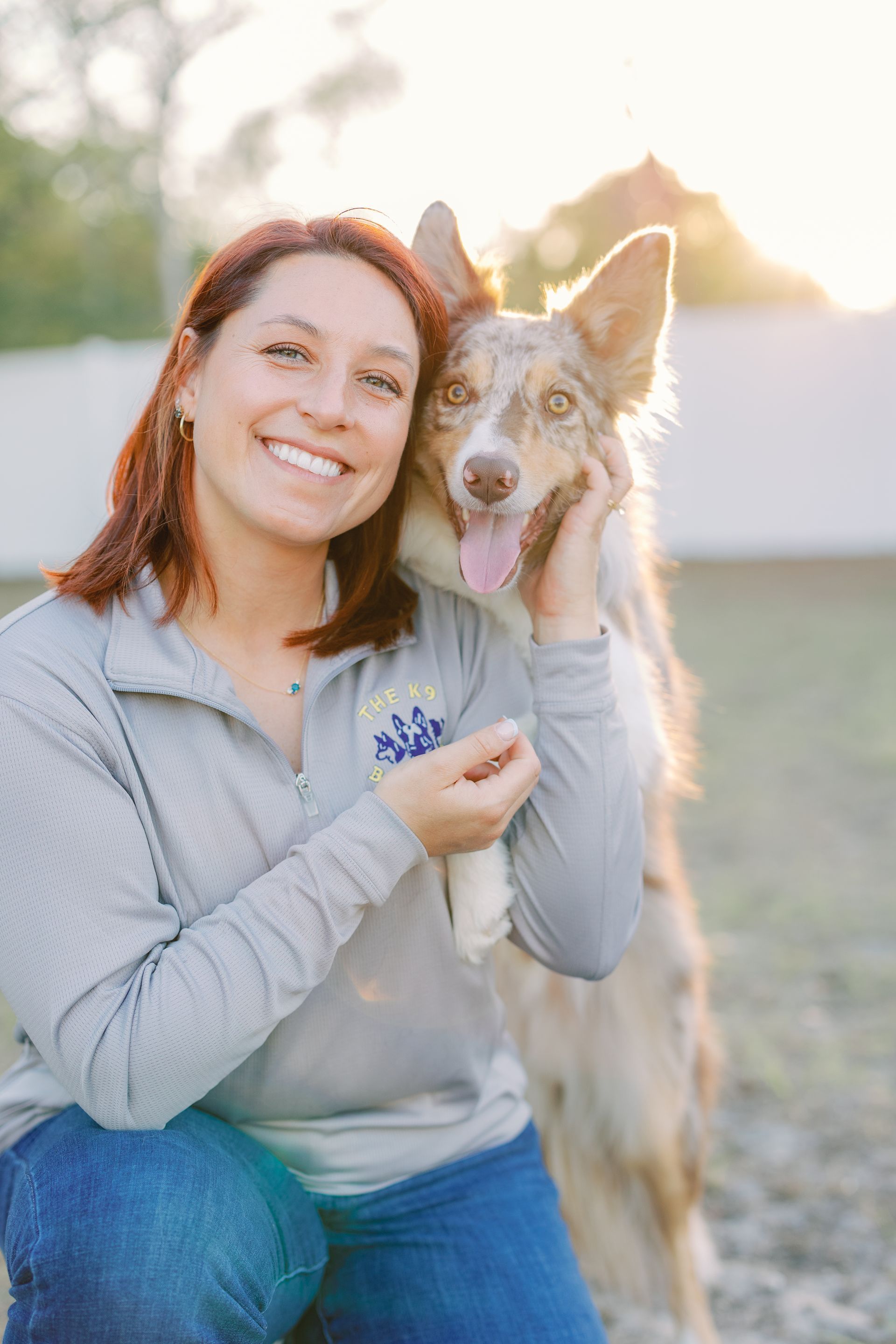 Woman kneeling outdoors and smiling beside a gray-brown dog at sunset