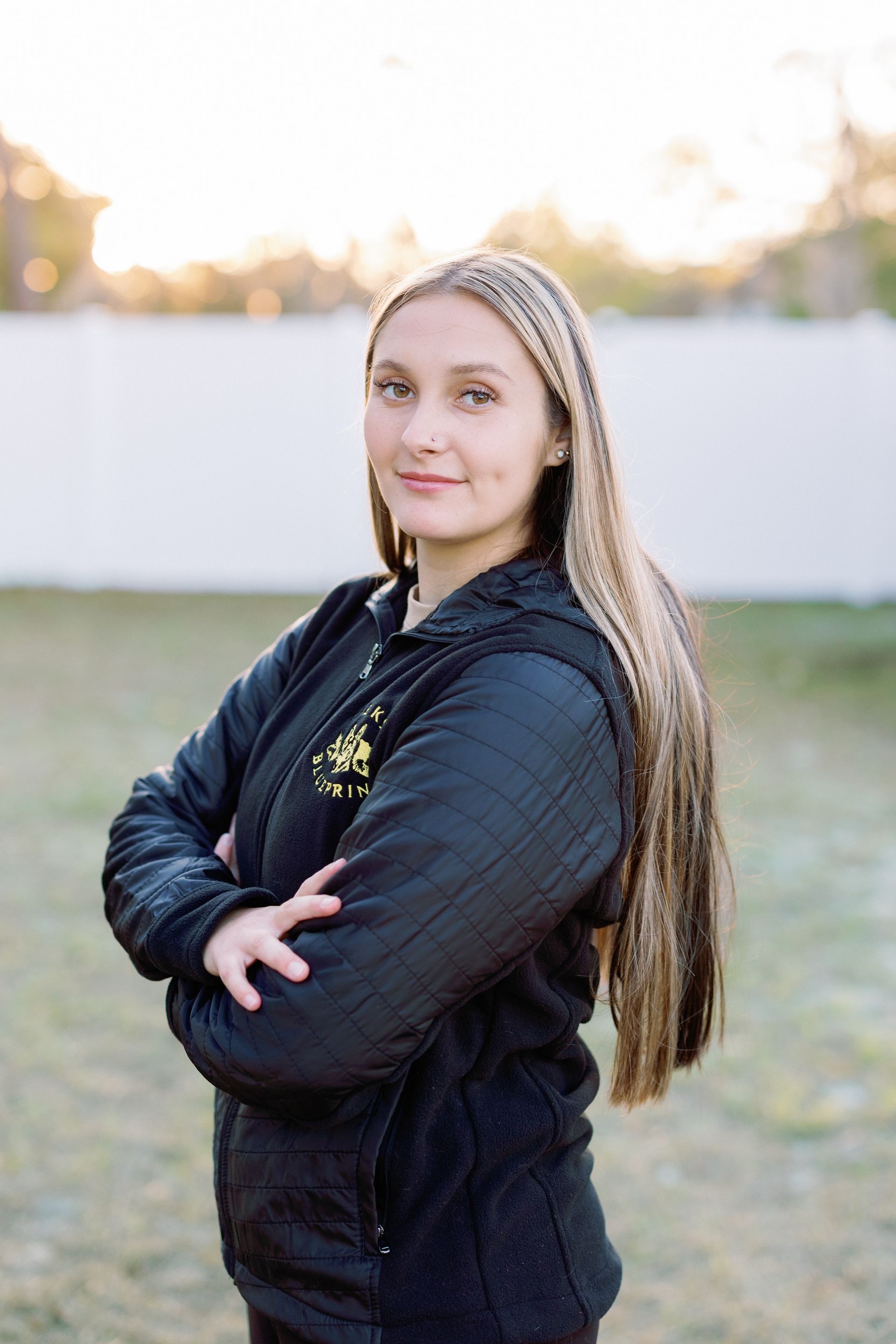 Woman with long braided hair stands with arms crossed outdoors in a black jacket, facing the camera.