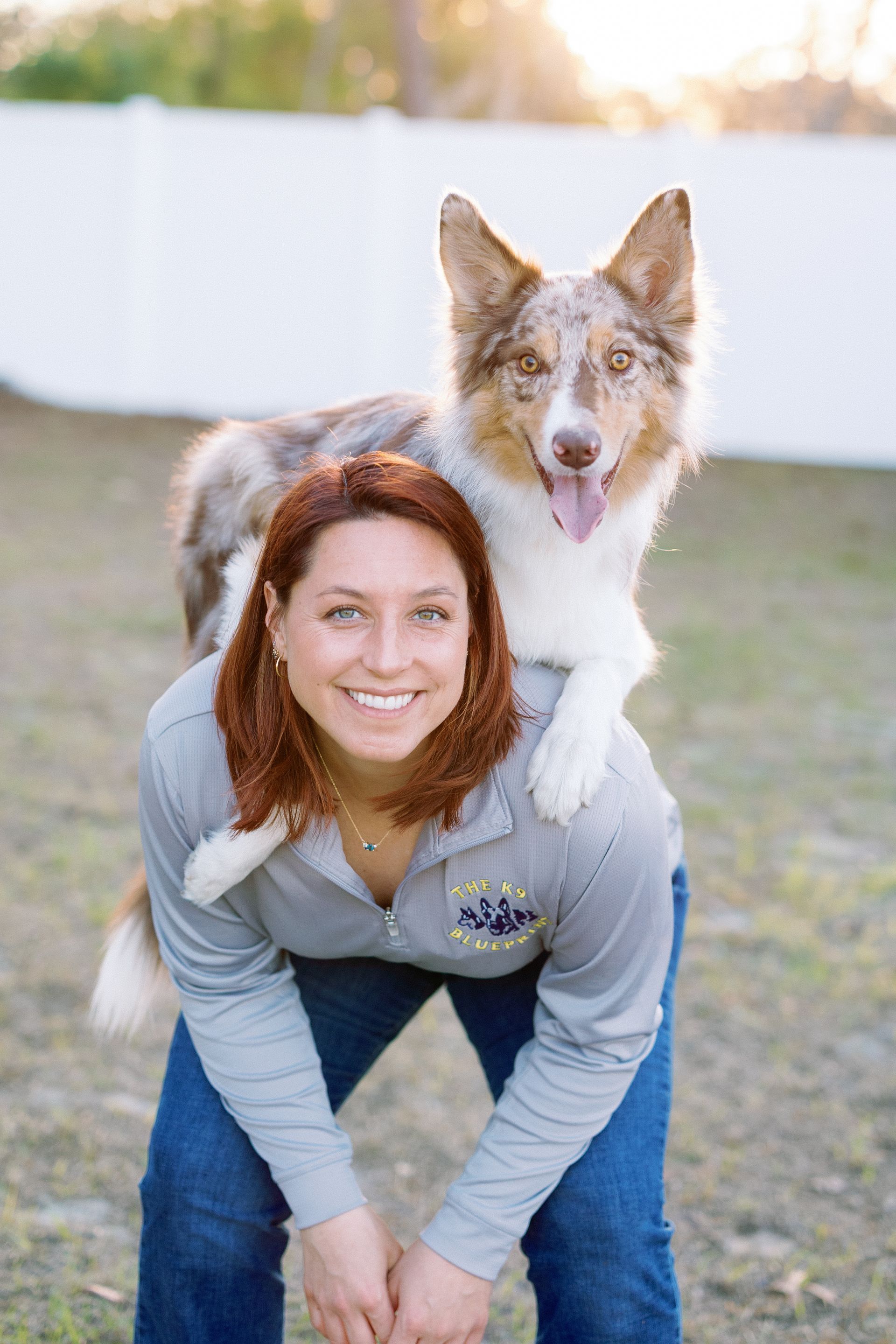 Woman crouches with brown-and-white dog on her shoulders in a grassy yard, both looking at camera.