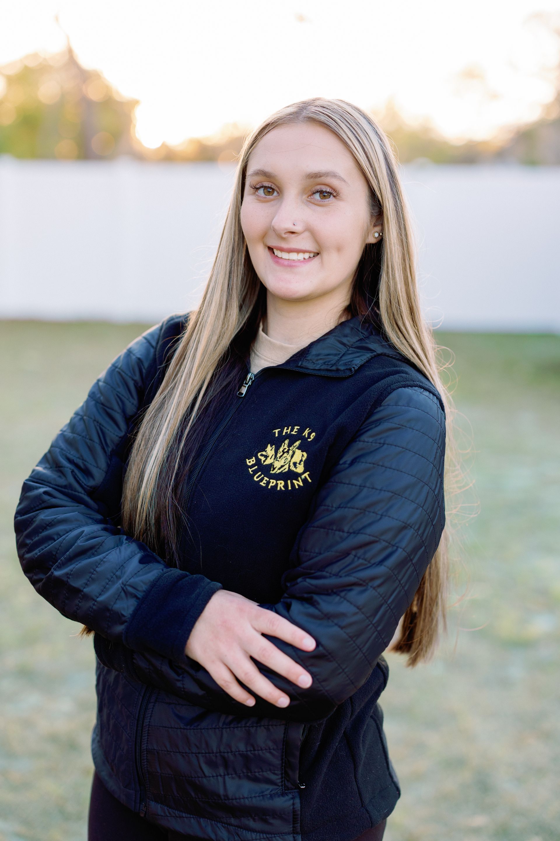 Smiling person in a black jacket standing outdoors with arms crossed
