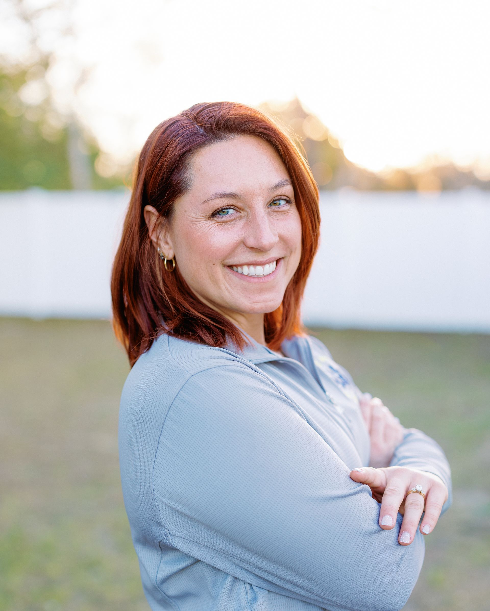 Smiling woman with red hair, arms crossed, standing outdoors in soft evening light.