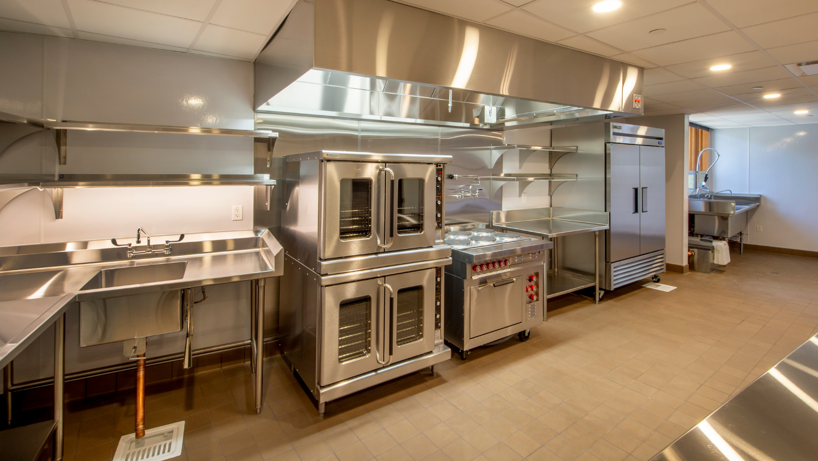 Commercial kitchen with stainless steel ovens, sinks, and counters under bright overhead lights.
