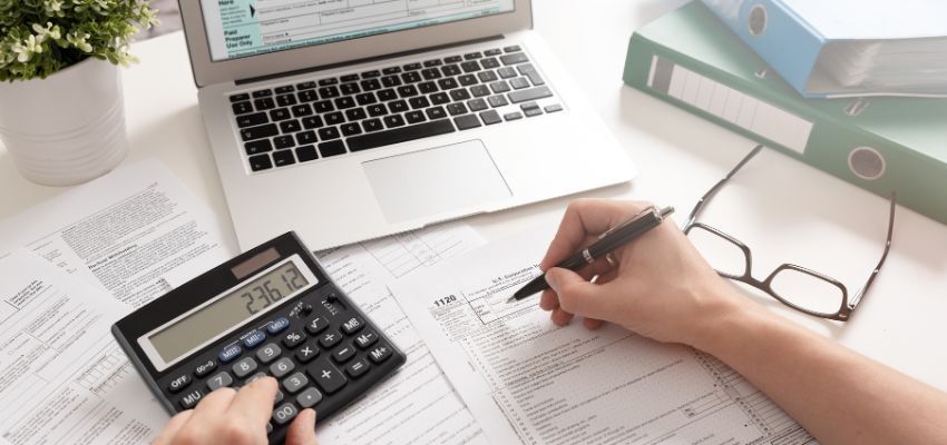 A hand writing on a tax form over a table with a laptop, calculator and files.