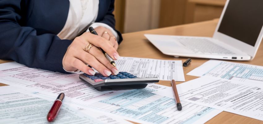 A hand writing on a tax form over a table with a laptop, calculator and files.