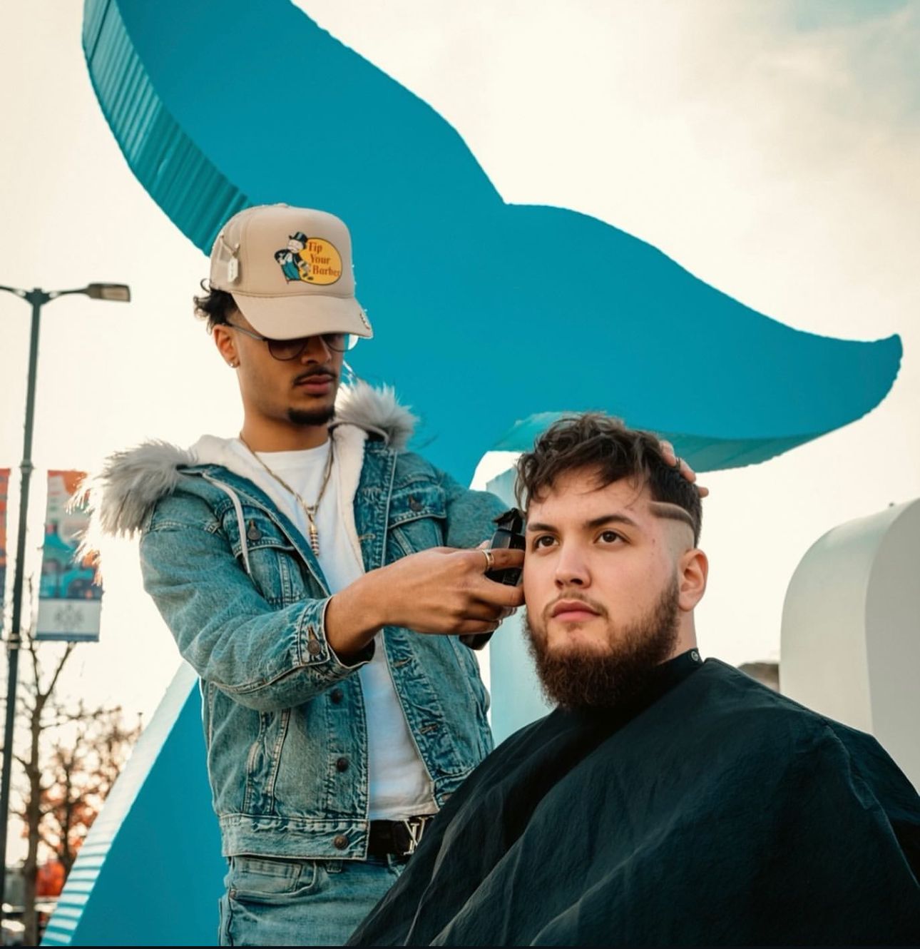 Barber giving a haircut outdoors, in front of a large blue whale tail sculpture.