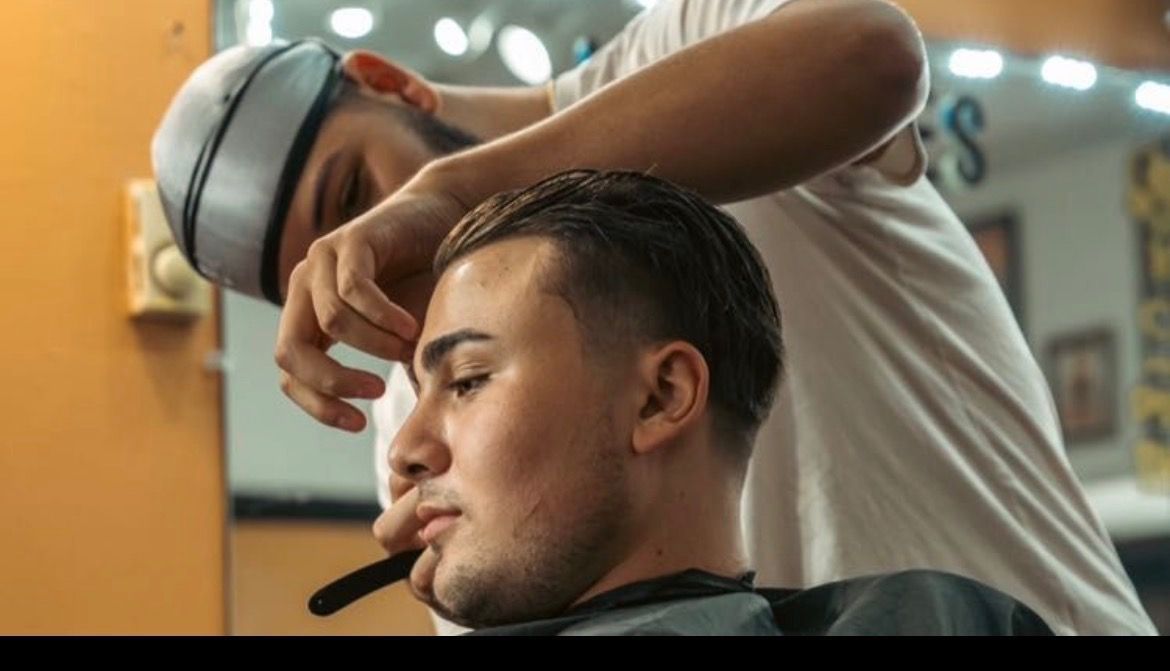 Barber cutting client's hair in a salon; client is seated and looking down, barber is standing with clippers.