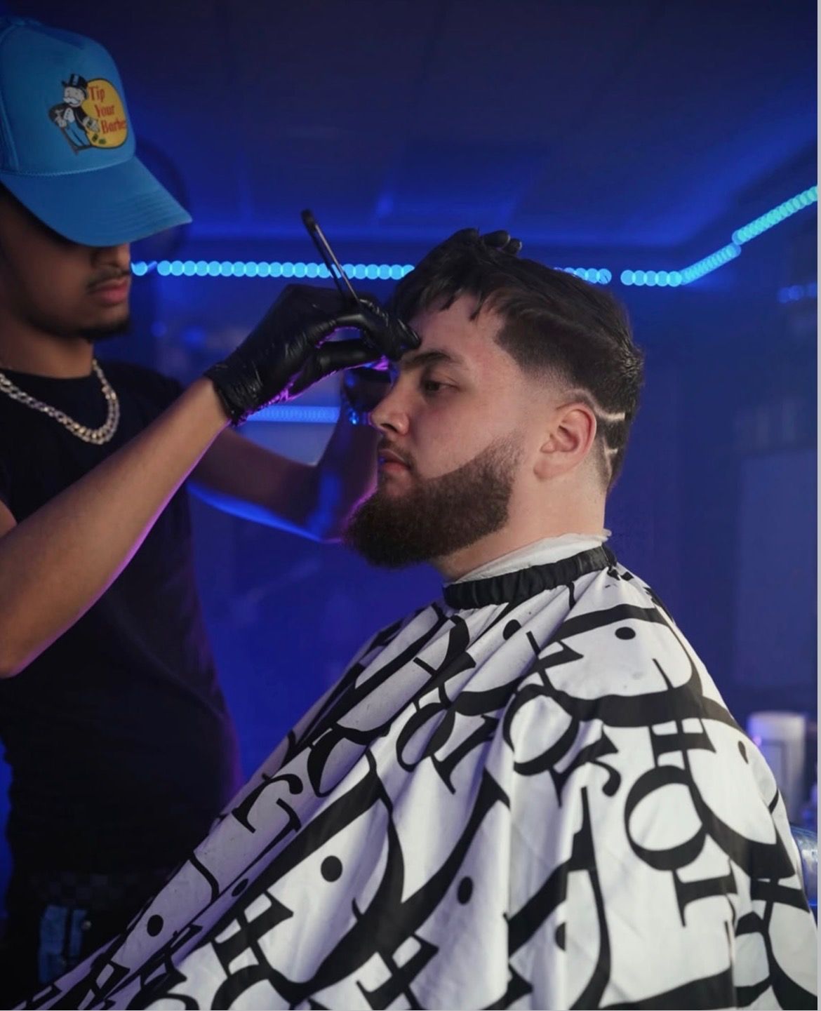 Barber cutting a client's hair, in a room with blue lights. Client wearing a Dior cape.