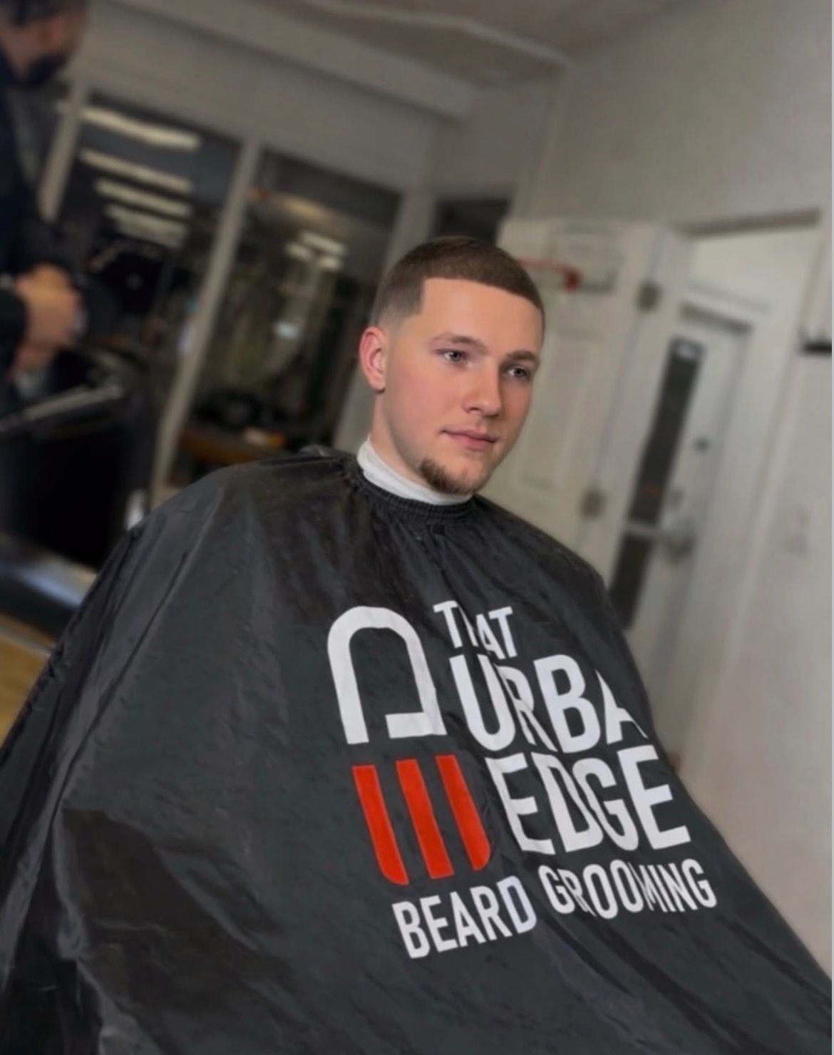 Man in barber's cape at "The Urban Edge Beard Grooming". Freshly cut hair, trimmed goatee, in a salon setting.