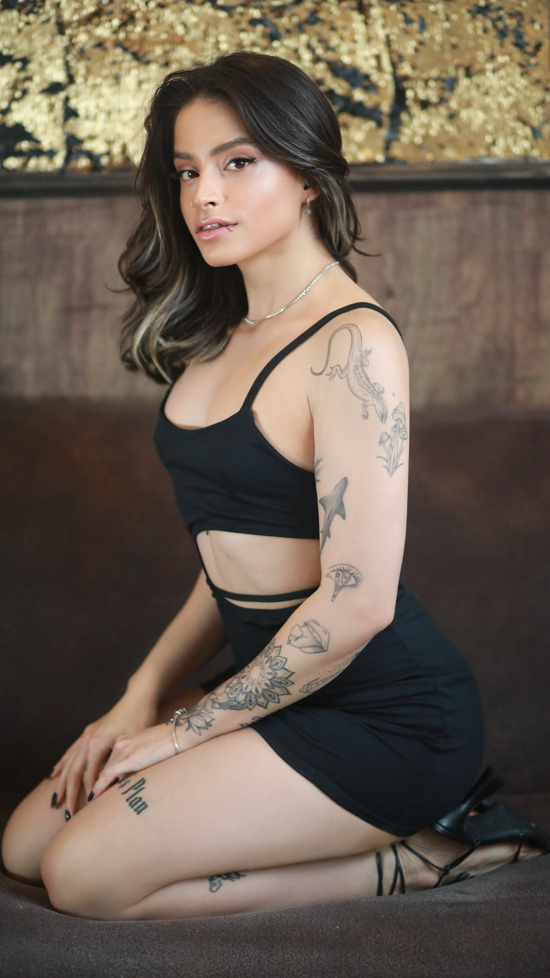 Uma pessoa com tatuagens nos braços ajoelha-se num sofá marrom, olhando para a câmera enquanto veste uma roupa preta.