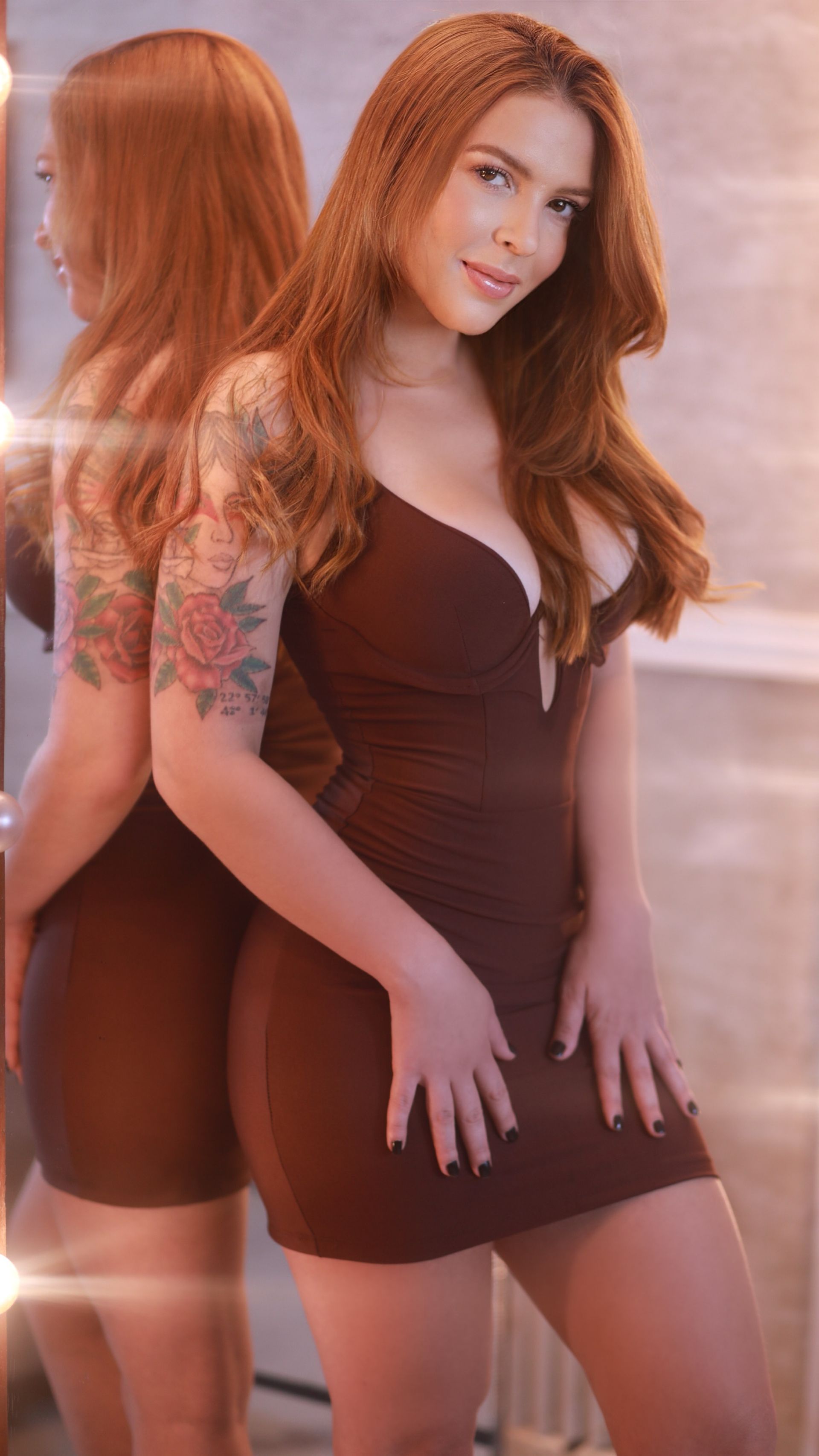 Uma pessoa sorridente, com longos cabelos castanho-avermelhados e uma grande tatuagem floral no braço.