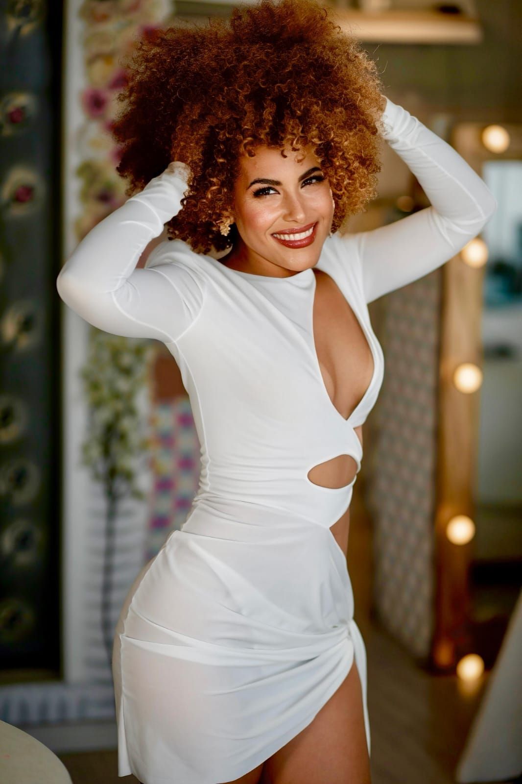 Uma pessoa com cabelo loiro trançado posa em uma escada, vestindo um vestido preto e sorrindo enquanto se apoia no corrimão.