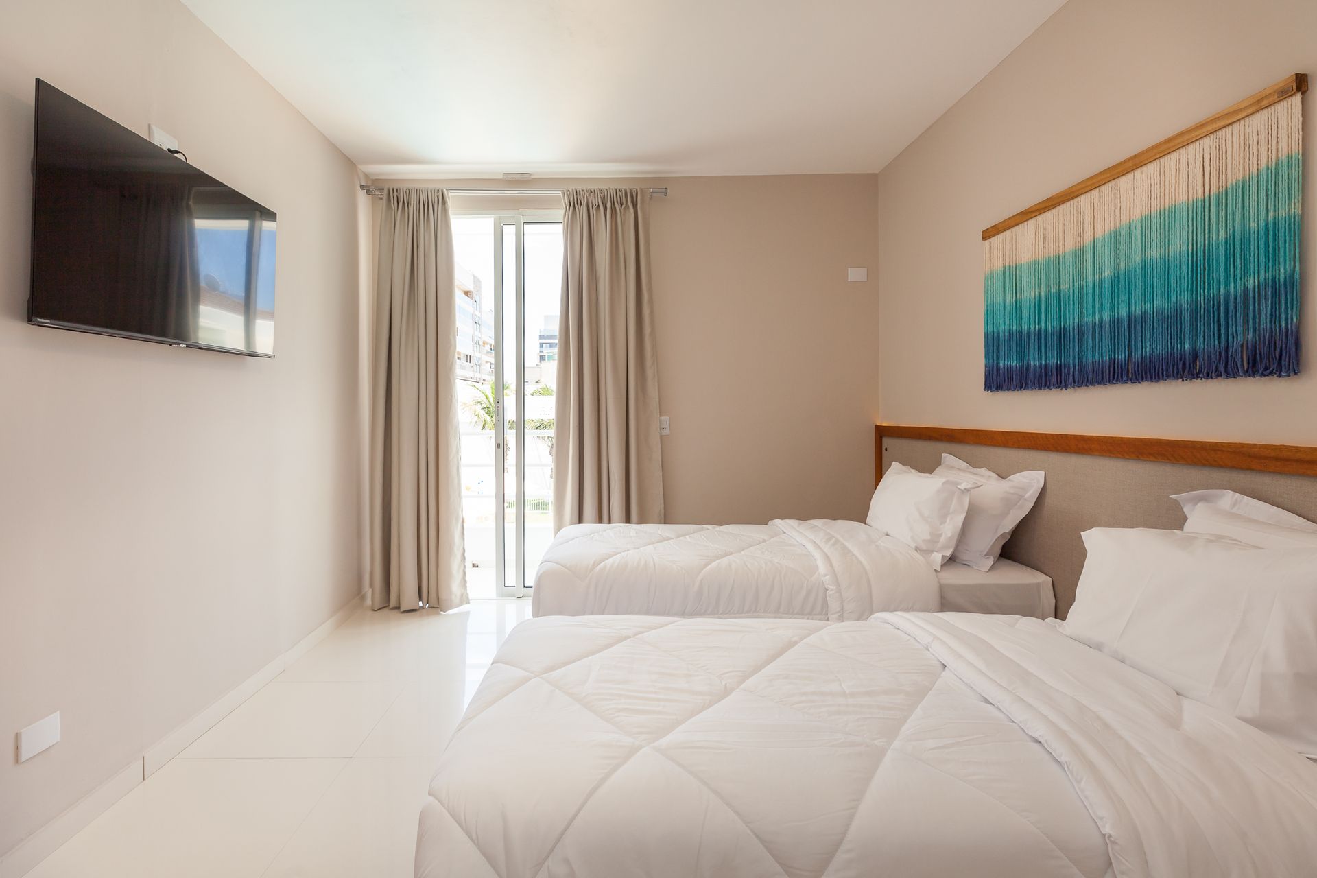 Quarto com duas camas de solteiro, TV na parede, portas para varanda e tapeçaria azul e branca na parede.