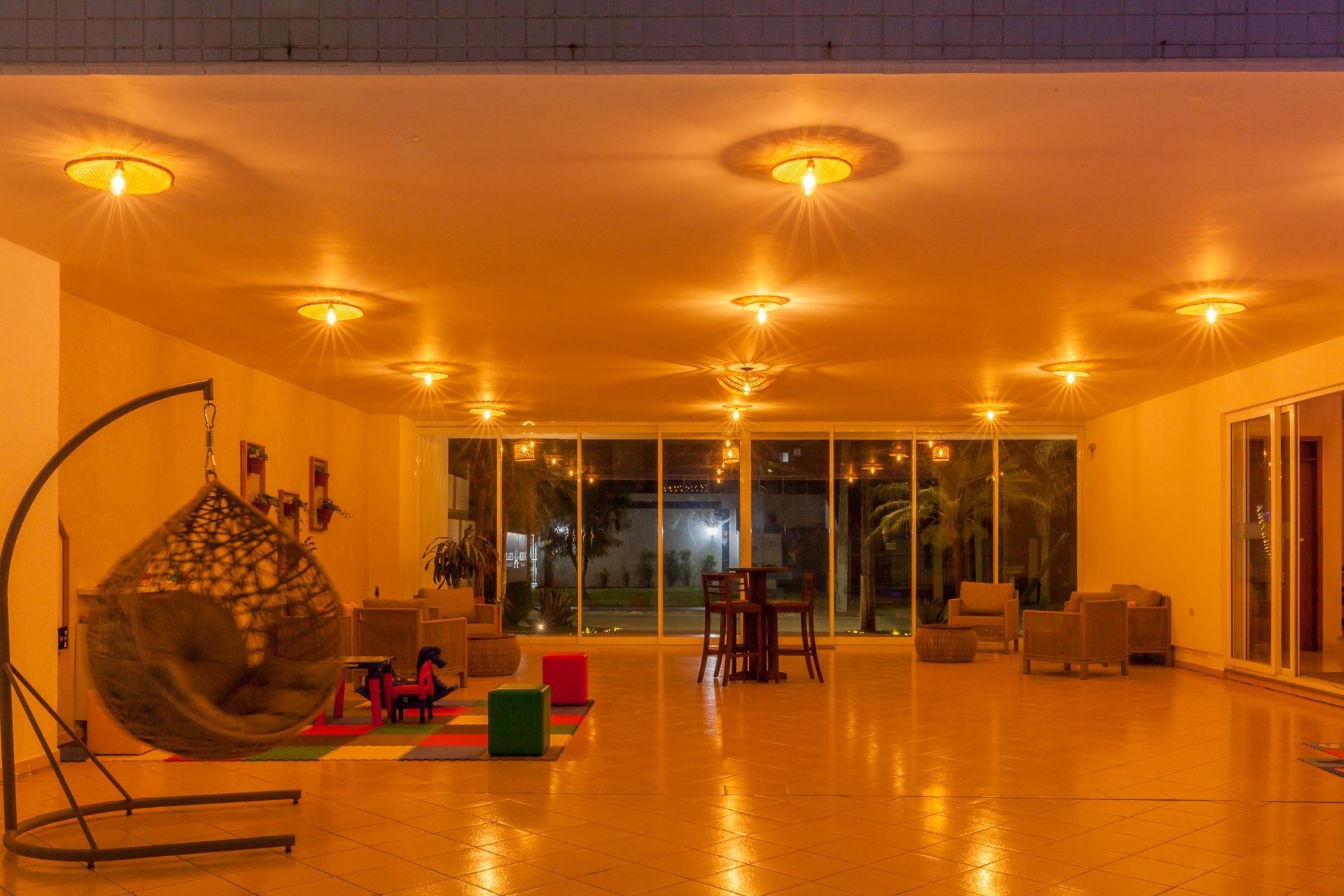 Quarto espaçoso com cadeira suspensa, iluminado por luzes de teto aconchegantes e com vista para uma área externa através de grandes janelas.