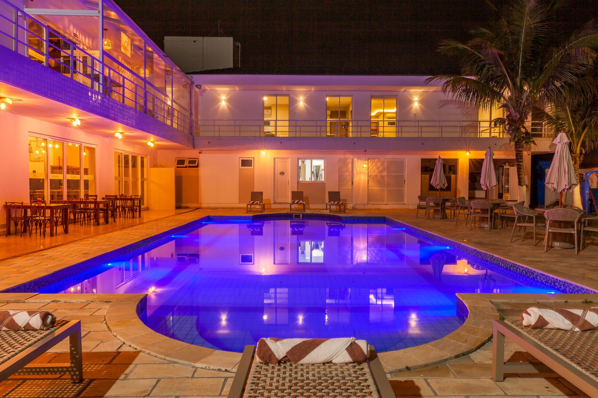 Vista noturna da piscina do hotel iluminada em azul, com cadeiras, mesas e o prédio.
