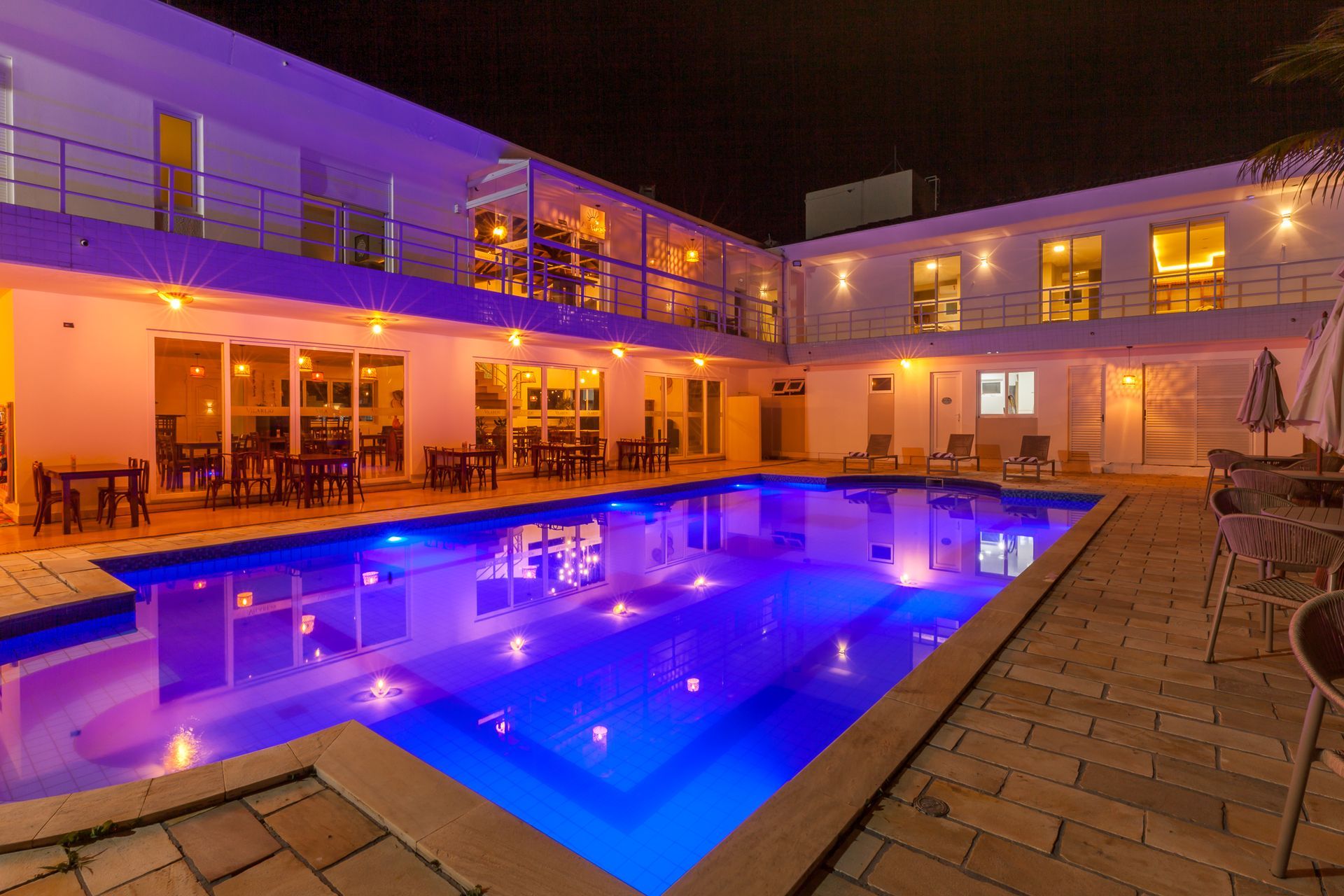 Pátio do hotel à noite, piscina iluminada, mesas e cadeiras, prédio de dois andares.
