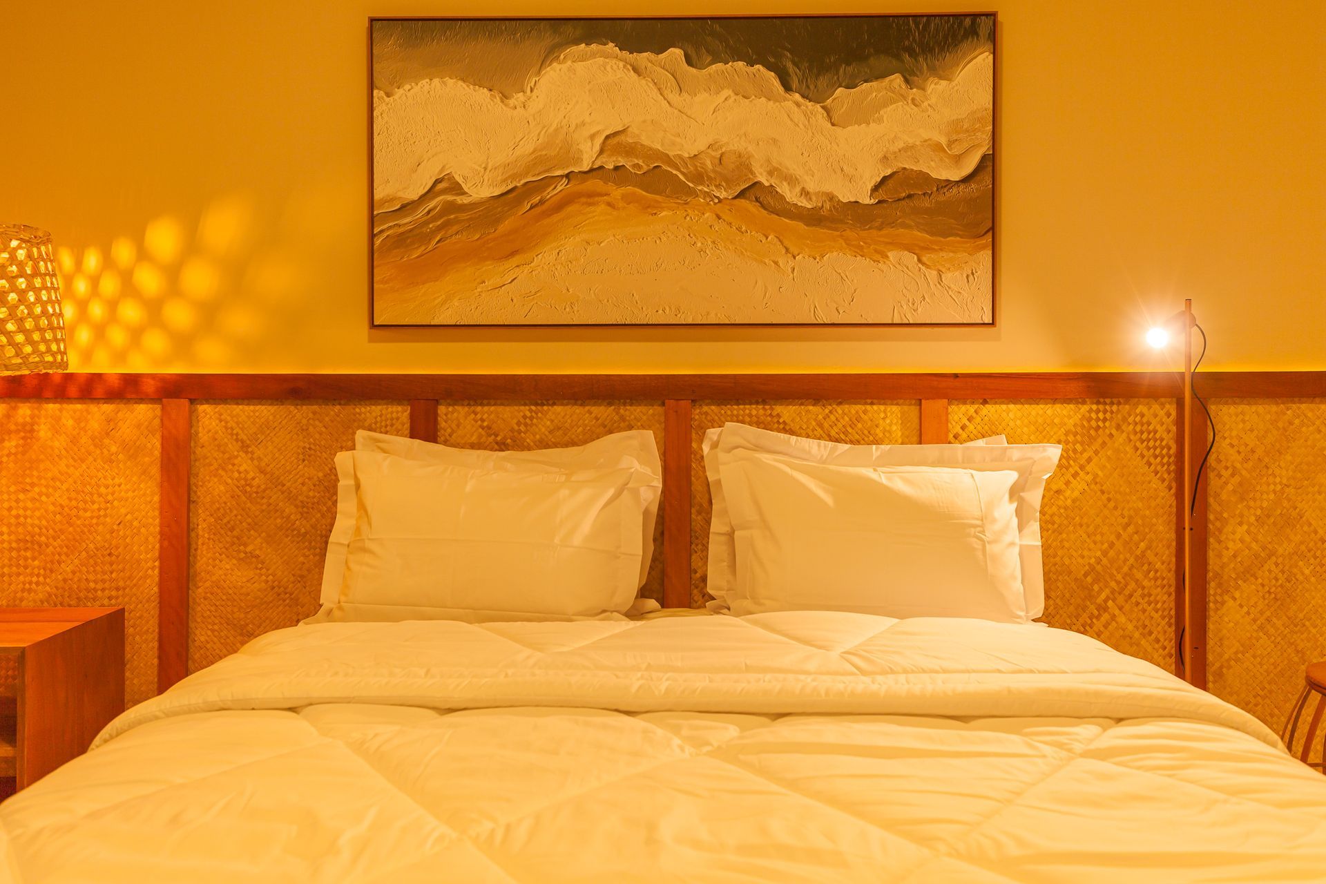 Quarto com cama, roupa de cama branca, cabeceira de vime e um quadro de ondas acima dela.
