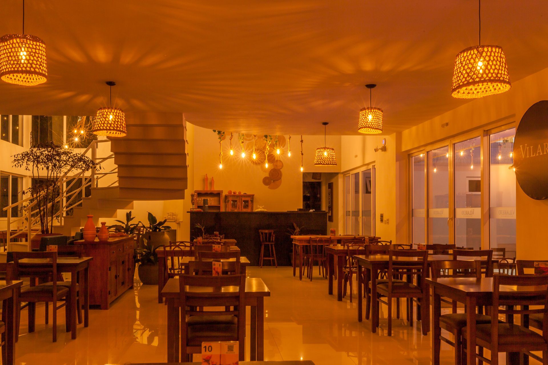 Interior de restaurante com mesas, cadeiras, bar e luminárias pendentes de madeira.
