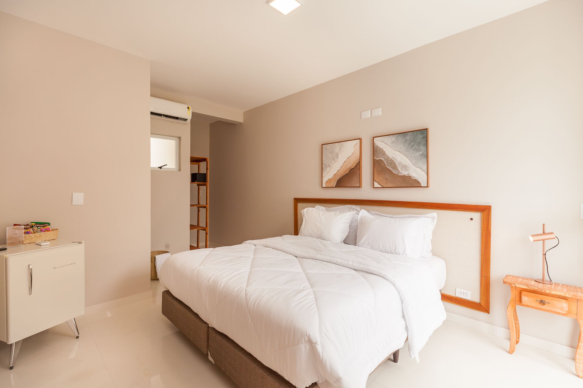 Quarto com cama branca, paredes bege e duas obras de arte abstratas na parede.