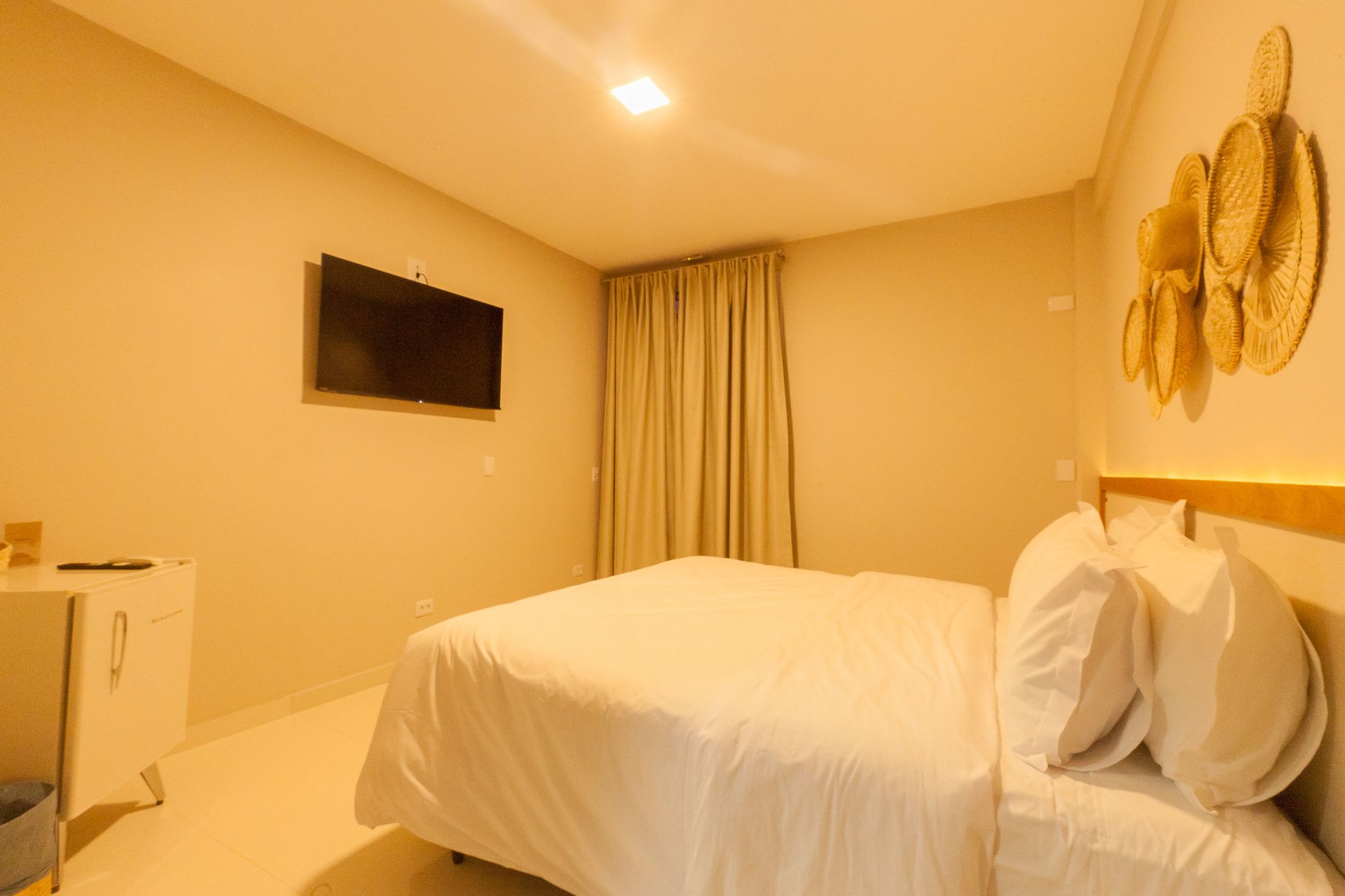 Quarto com cama, TV, cortinas e decoração de parede. O quarto é bem iluminado e tem paredes brancas.