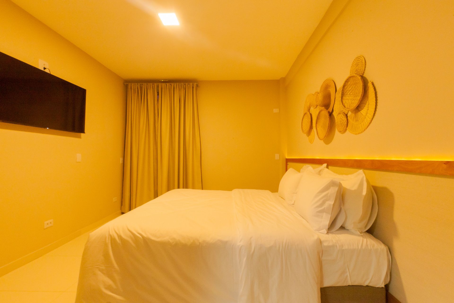 Quarto com cama, TV, cortinas e decoração de parede; paredes e iluminação amarelas.