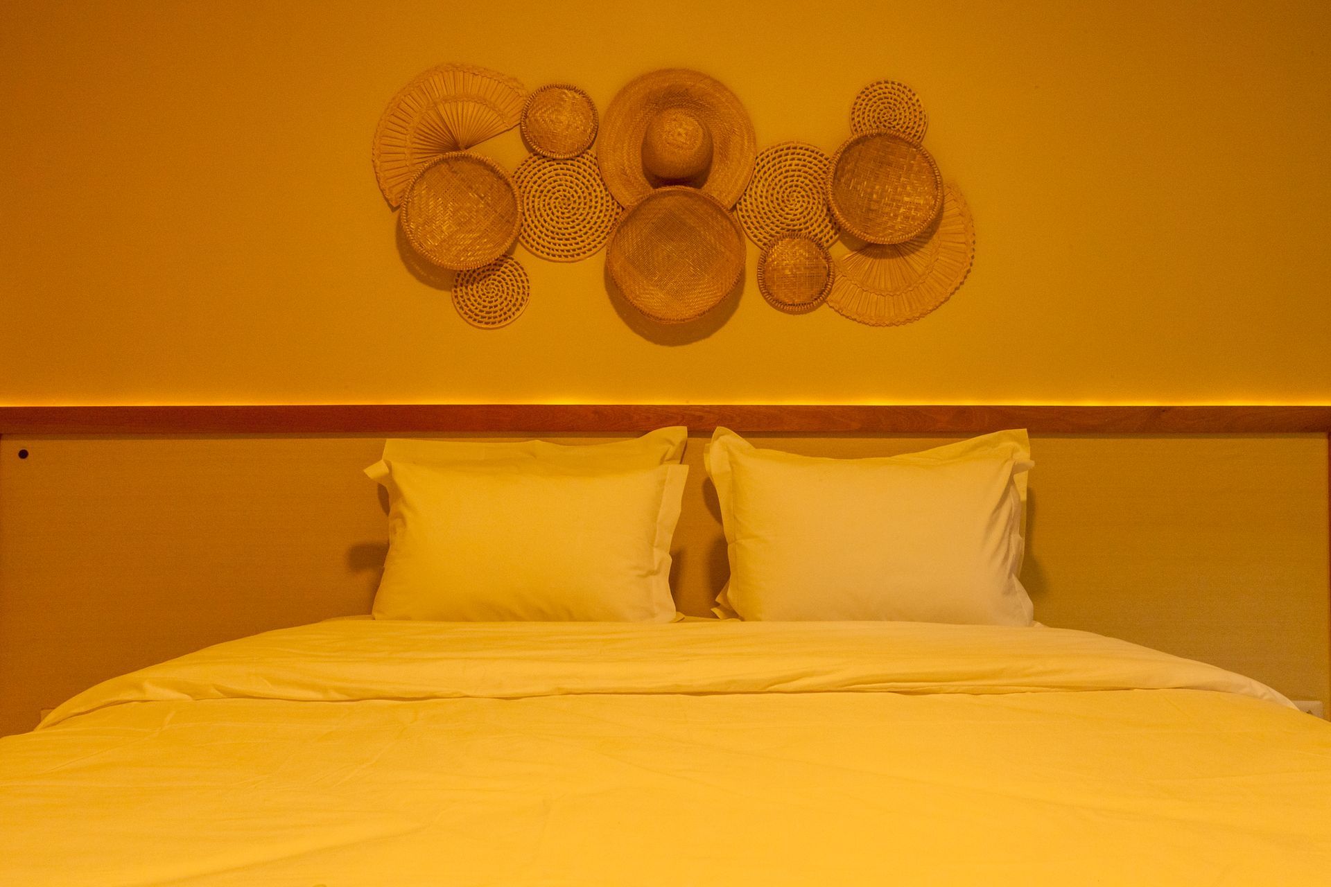 Cama com travesseiros e roupa de cama brancos, sob uma obra de arte em palha na parede, em um quarto com iluminação amarela.