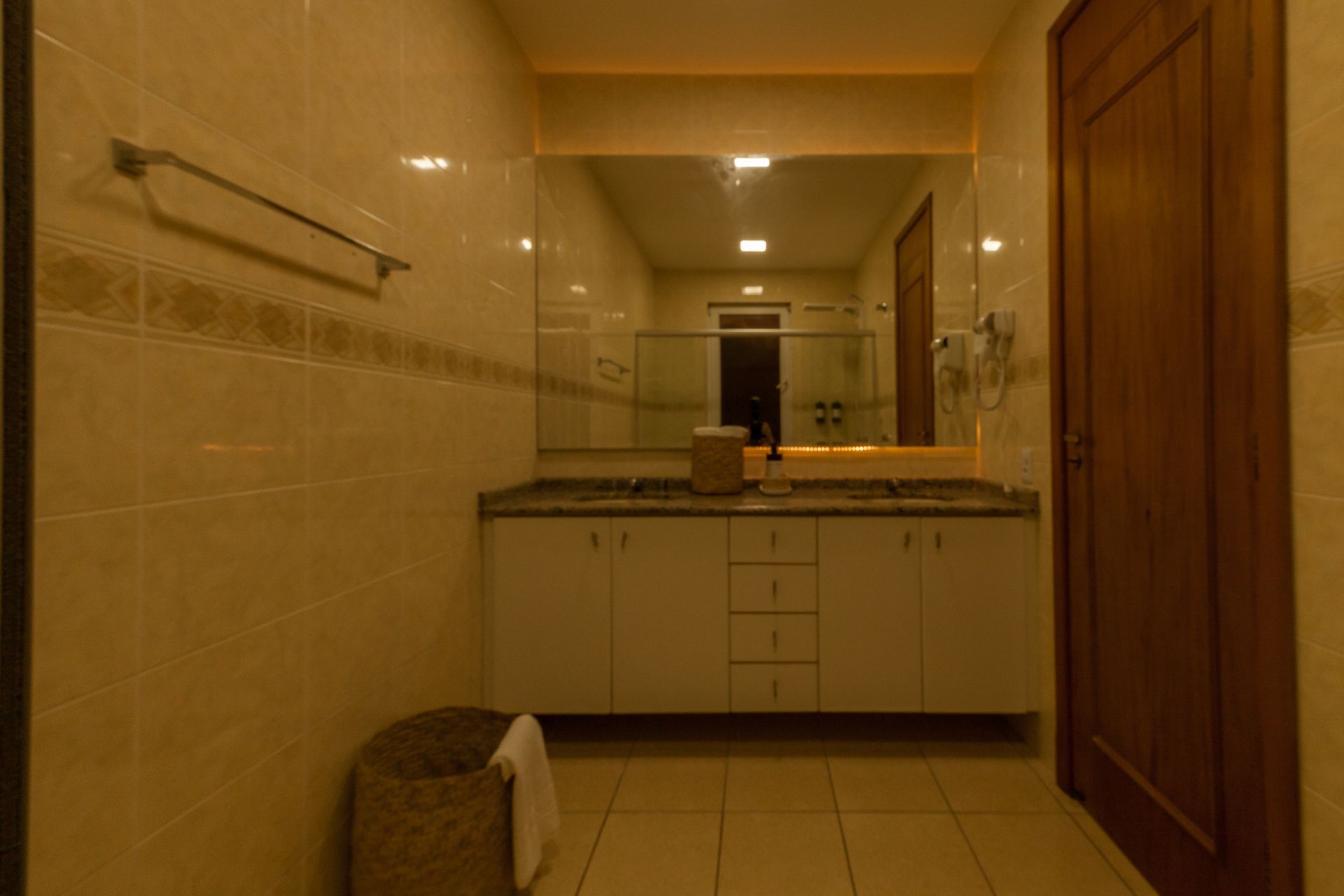 Banheiro com paredes revestidas de azulejos cor creme, um grande espelho, armários brancos e uma porta de madeira.