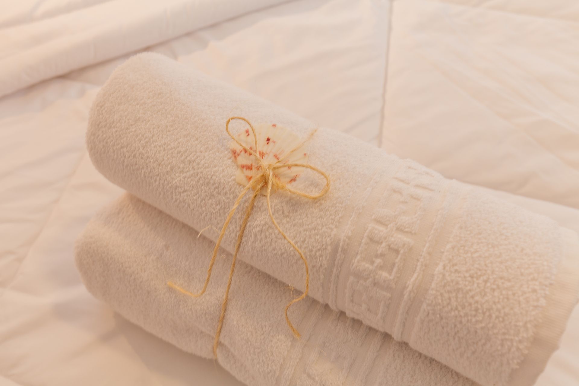 Toalhas brancas enroladas, amarradas com barbante e uma flor decorativa, sobre uma cama branca.