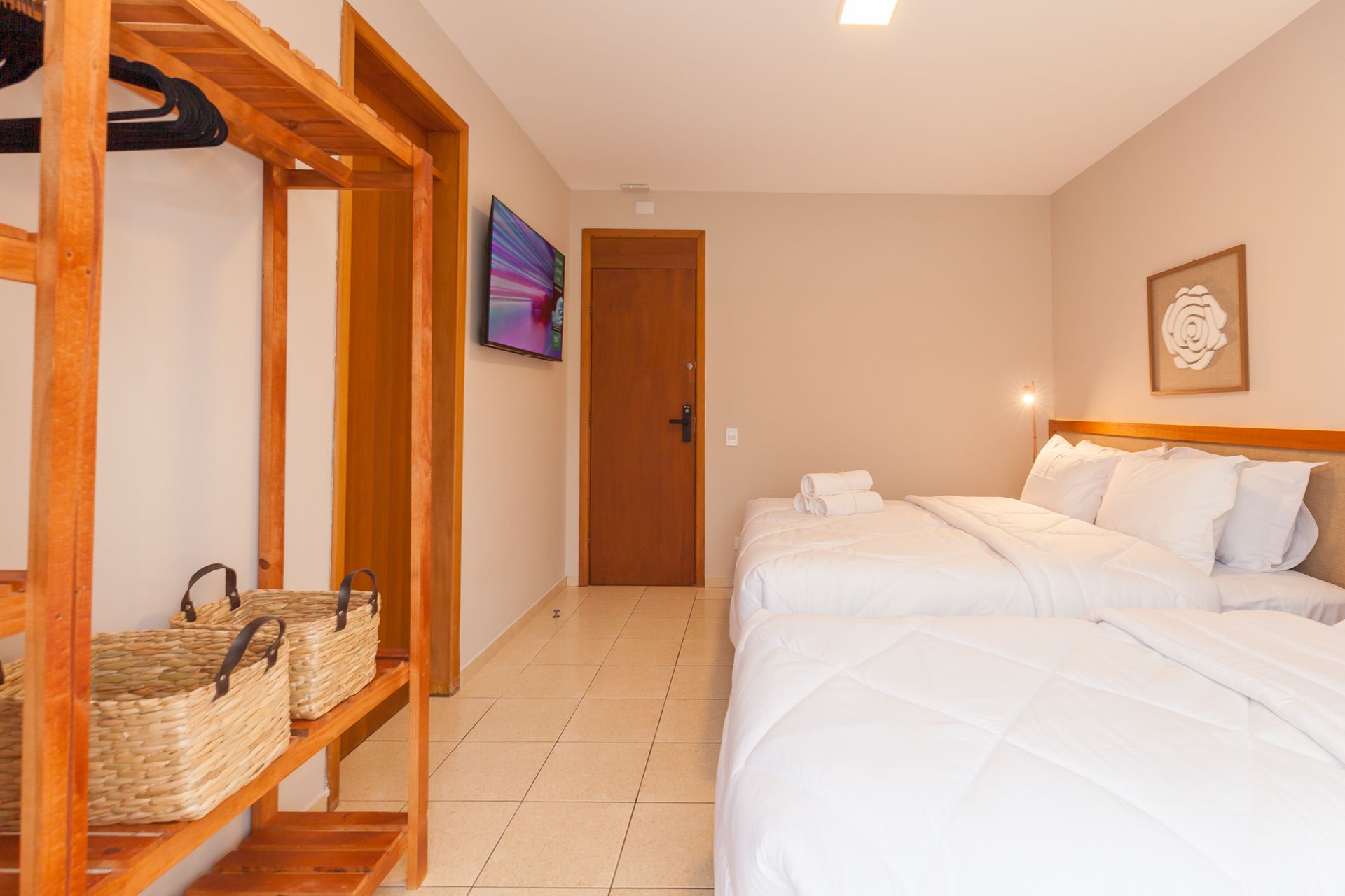 Quarto com duas camas, armário aberto e TV na parede. Paredes bege, detalhes em madeira e piso de azulejo.