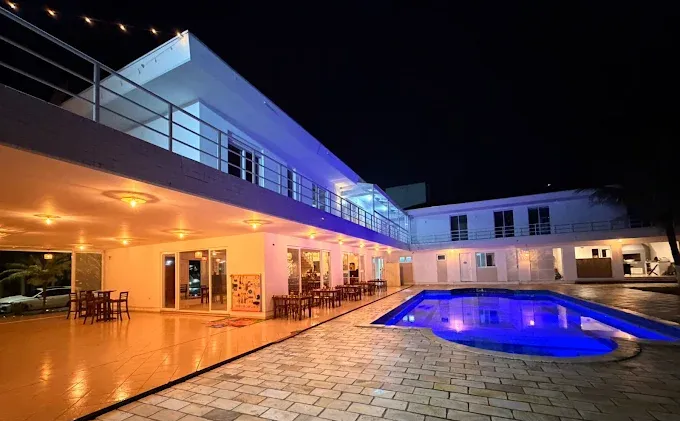 Vista noturna de um hotel moderno com piscina. O edifício é branco, com iluminação azul. Mesas e cadeiras estão dispostas em um pátio.