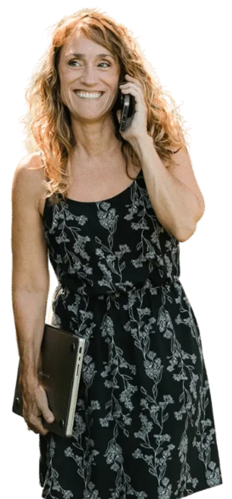 A woman in a black dress is holding a laptop and talking on a cell phone.