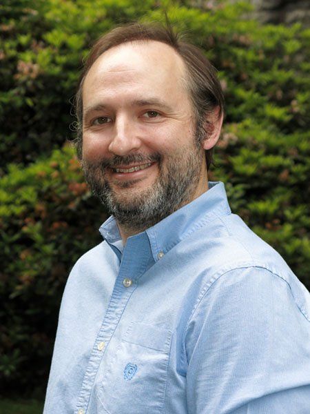 A smiling person with a beard, wearing a light blue button-down shirt, standing in front of green shrubbery.