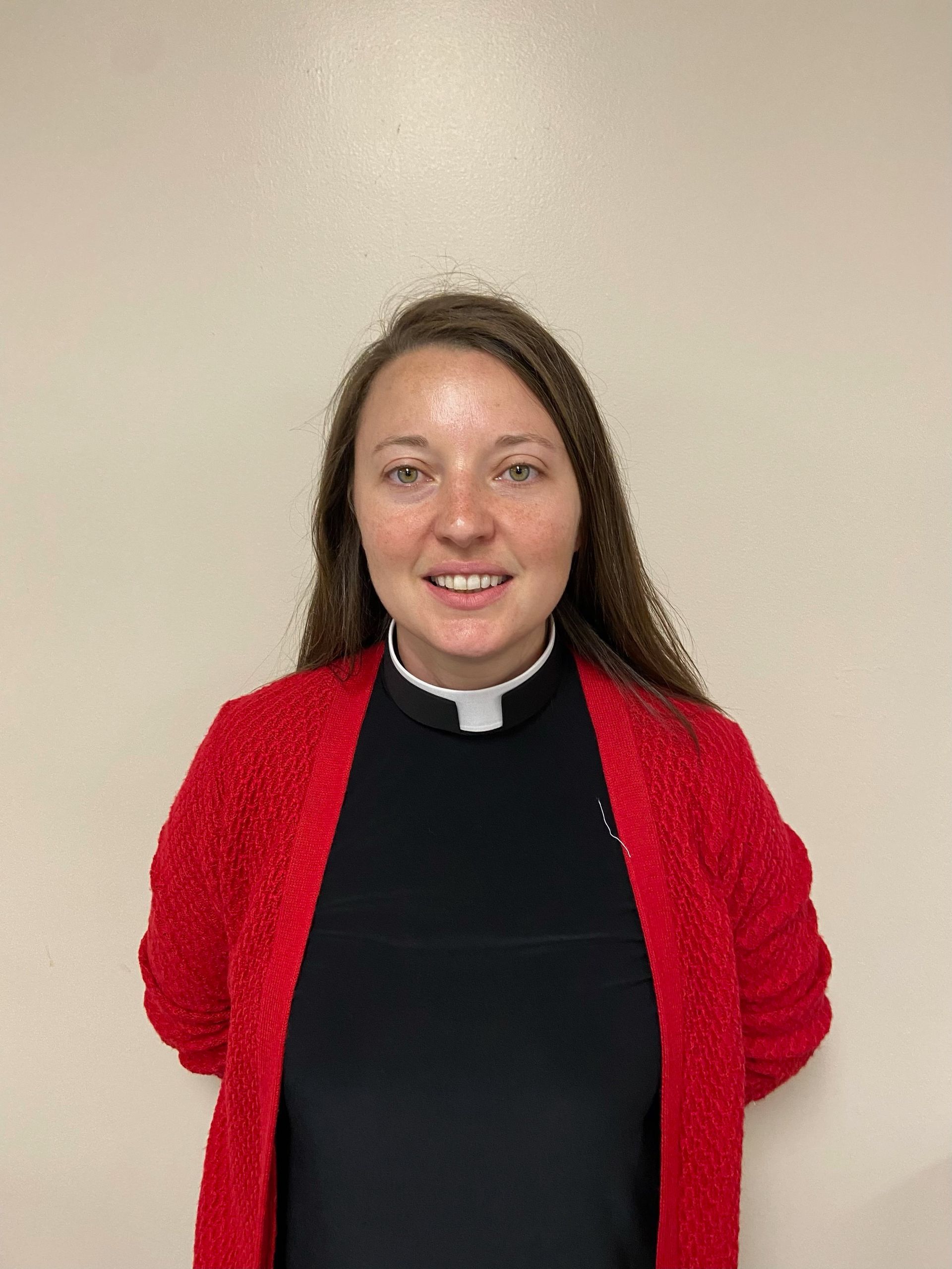 A smiling individual wearing a black clerical shirt with a white tab collar under a bright red, textured cardigan.