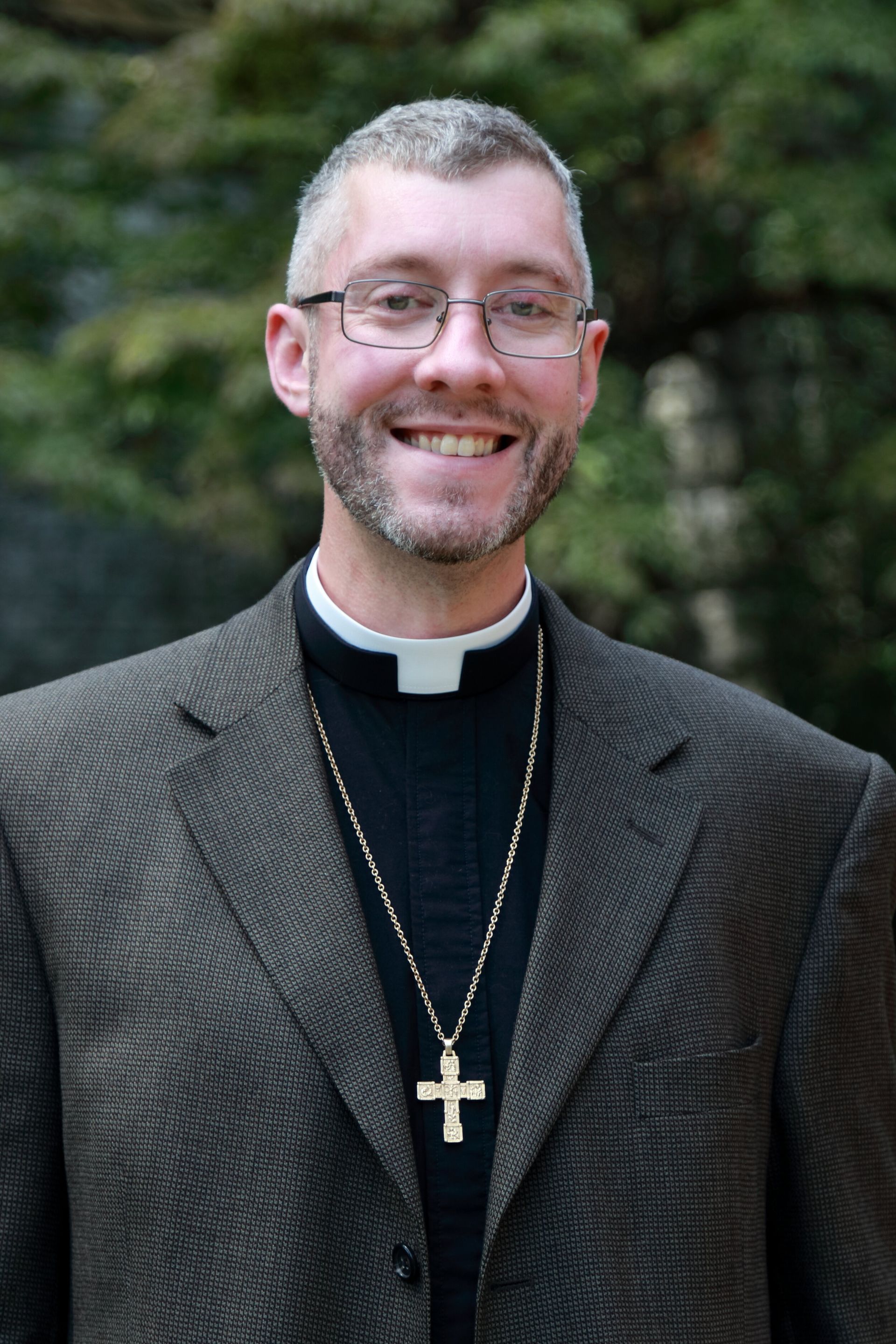 A person wearing glasses, a dark suit, and a clerical collar with a cross necklace, smiling against a blurred green background.
