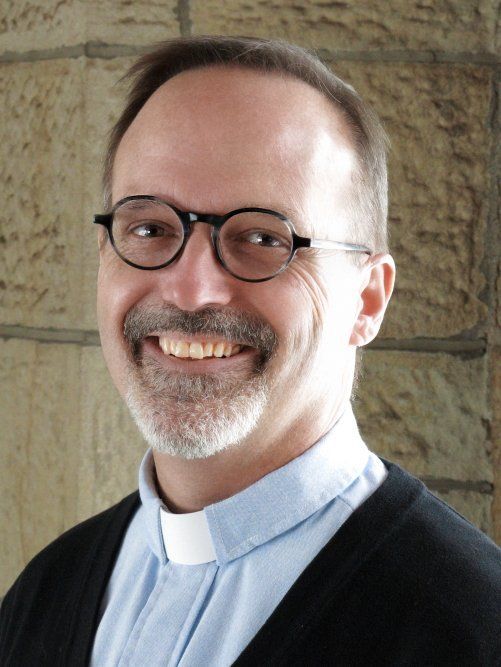 A smiling person wearing glasses, a light blue shirt with a clerical collar, and a black sweater against a stone wall.