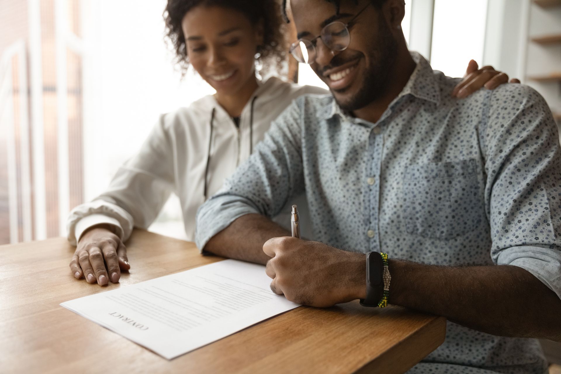 Two people reviewing and signing a document at a wooden table near a window.