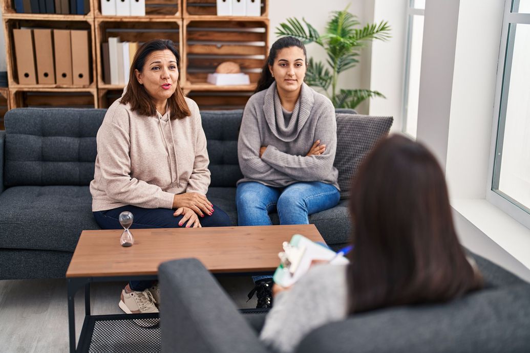 Two women sitting on a couch in a counseling session, facing a therapist across a coffee table.