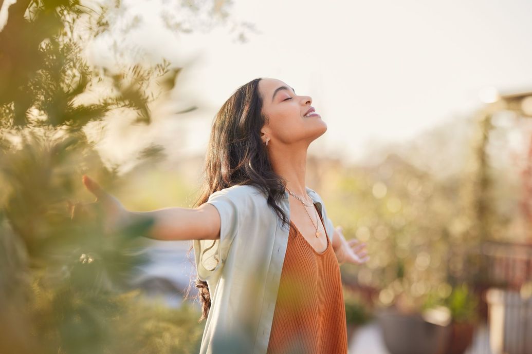 Woman with arms outstretched in a sunlit garden, looking up with a peaceful expression.