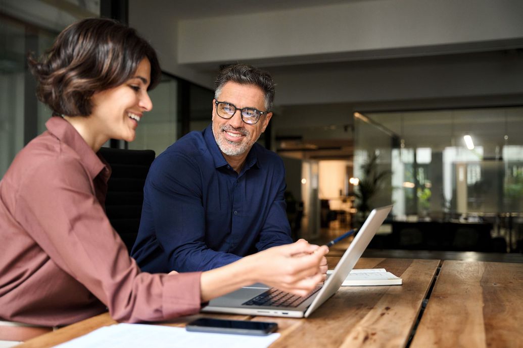 Two coworkers smiling at a laptop across a wooden table in an office